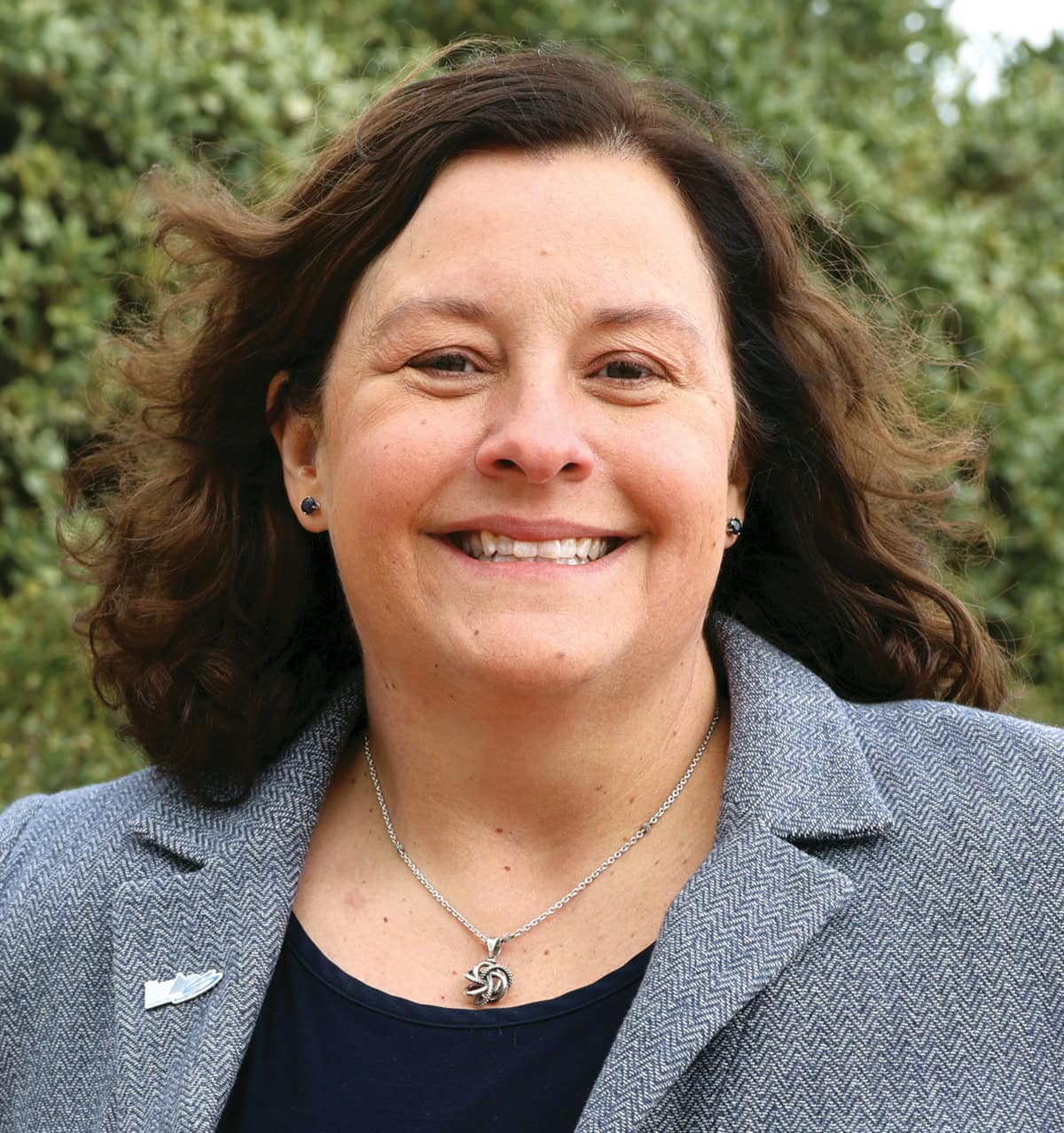 Andrea Ross smiling, wearing a blazer and necklace, outdoors with green foliage in the background.